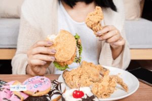 Woman holding burger and fried chicken with desserts on table, symbolizing binge eating disorder treatment and recovery at Empower Health Group