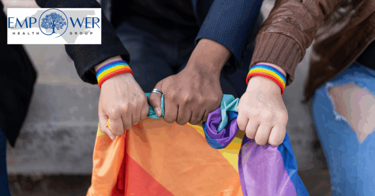 A close-up of three people sitting side by side, each wearing a rainbow wristband and holding a folded Pride flag together. The image reflects unity and support, with the Empower Health Group logo displayed in the upper left corner.