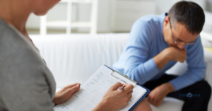 A therapist in the foreground takes notes during a session with a man on a white couch.